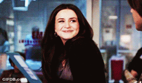 A Woman In A Black Sweater Is Smiling And Looking At The Camera In A Room GIF