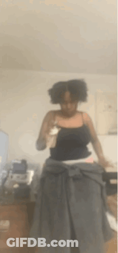 A Woman In A Black Tank Top Is Standing In A Living Room Holding Her Hair . GIF