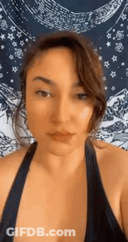 A Woman In A Black Tank Top Is Standing In Front Of A Tapestry And Looking At The Camera . GIF