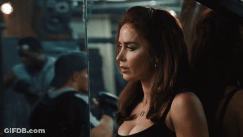 A Woman In A Black Tank Top Is Standing In Front Of A Window GIF