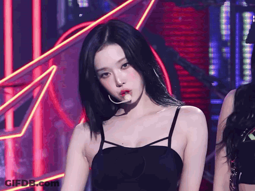 A Woman In A Black Tank Top With A Microphone In Her Mouth Looks At The Camera GIF