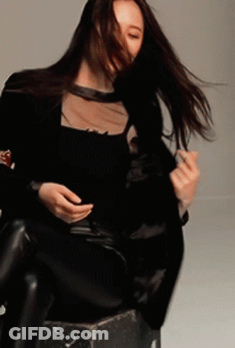 A Woman In A Black Top And Black Pants Is Sitting On A Stool With Her Hair Blowing In The Wind GIF