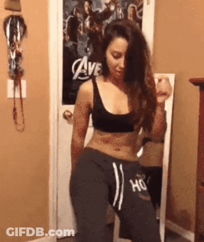 A Woman In A Black Top And Grey Sweatpants Is Dancing In Front Of A Mirror In A Room . GIF