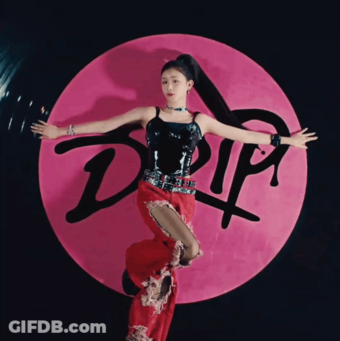 A Woman In A Black Top And Red Pants Stands In Front Of A Pink Circle That Says Drip GIF