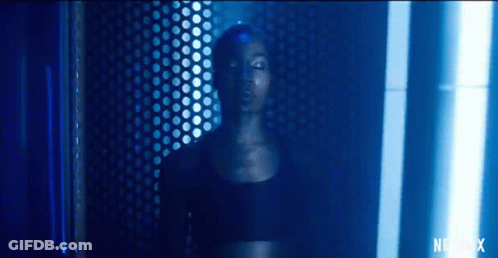 A Woman In A Black Top Is Standing In A Dark Room With A Blue Light Shining On Her Face . GIF