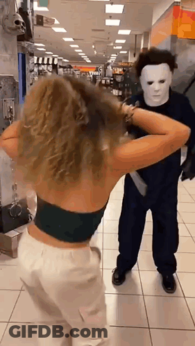 A Woman In A Black Top Is Standing Next To A Man In A Mask In A Store . GIF
