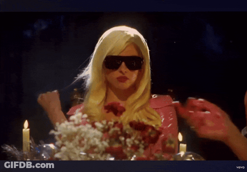 A Woman In A Blonde Wig And Sunglasses Is Sitting At A Table With Candles And Flowers GIF