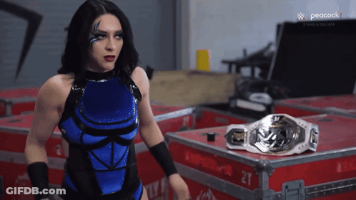 A Woman In A Blue And Black Wrestling Outfit Stands In Front Of A Peacock Logo GIF