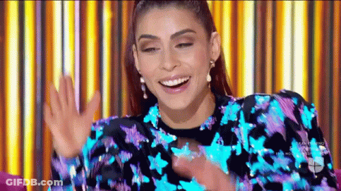 A Woman In A Blue And Purple Sequined Dress Is Smiling And Waving Her Hands . GIF