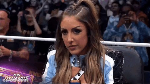 A Woman In A Blue And White Jacket Is Sitting In A Wrestling Ring In Front Of A Crowd . GIF