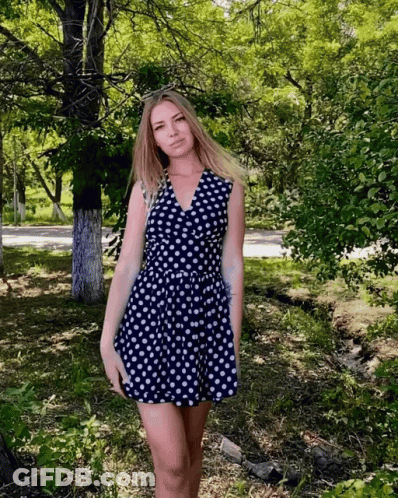 A Woman In A Blue And White Polka Dot Dress Stands In A Park GIF