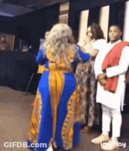 A Woman In A Blue And Yellow Dress Is Dancing In Front Of A Crowd Of People . GIF