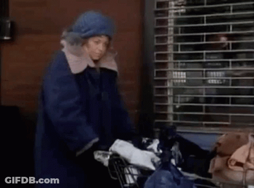 A Woman In A Blue Coat Is Pushing A Shopping Cart With A Bag Of Trash . GIF