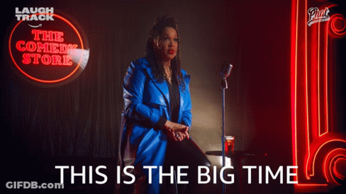 A Woman In A Blue Coat Is Sitting In Front Of A Neon Sign That Says The Comedy Store GIF