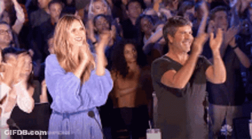 A Woman In A Blue Dress And A Man In A Black Shirt Are Applauding In Front Of A Crowd . GIF