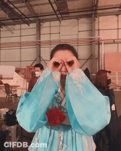 A Woman In A Blue Dress Covering Her Eyes With Her Hands In A Warehouse GIF