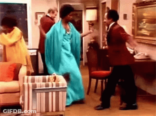 A Woman In A Blue Dress Is Dancing In A Living Room With A Man In A Red Suit . GIF