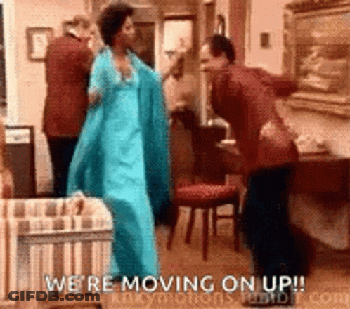 A Woman In A Blue Dress Is Dancing In A Living Room With A Man In A Suit . GIF