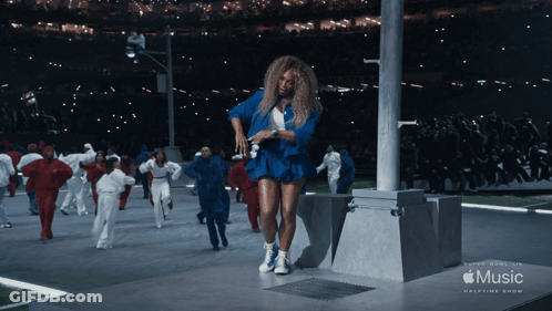 A Woman In A Blue Dress Is Dancing In Front Of A Music Halftime Show Banner GIF