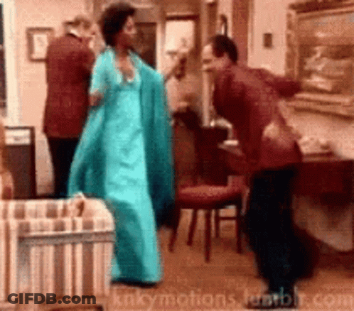 A Woman In A Blue Dress Is Dancing With A Man In A Red Suit In A Living Room GIF