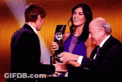 A Woman In A Blue Dress Is Holding A Trophy And Shaking Hands With A Man In A Suit GIF