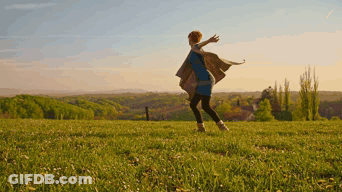 A Woman In A Blue Dress Is Standing In A Grassy Field With Her Arms Outstretched GIF