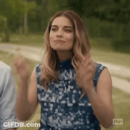 A Woman In A Blue Dress Is Standing In A Park With Her Hands In The Air . GIF
