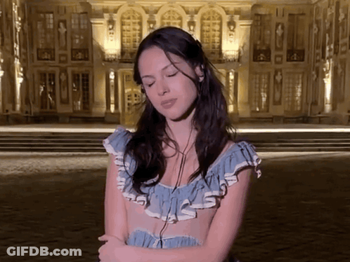 A Woman In A Blue Dress Is Standing In Front Of A Building With Her Eyes Closed GIF