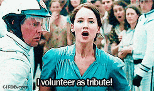 A Woman In A Blue Dress Is Standing In Front Of A Crowd Of People And Says `` I Volunteer As Tribute ! '' GIF