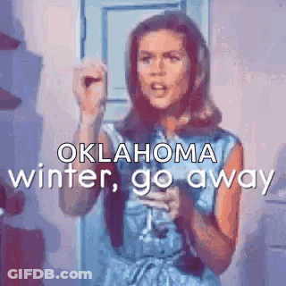 A Woman In A Blue Dress Is Standing In Front Of A Door Holding A Microphone And Saying Oklahoma Winter Go Away . GIF