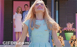 A Woman In A Blue Dress Is Standing In Front Of A Pink House With Her Arms Outstretched . GIF