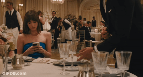 A Woman In A Blue Dress Sits At A Table Looking At Her Cell Phone GIF