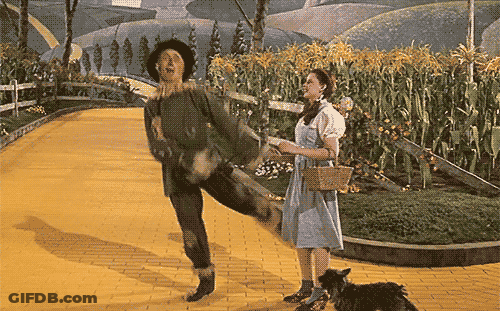 A Woman In A Blue Dress Stands Next To A Scarecrow On A Yellow Brick Road GIF