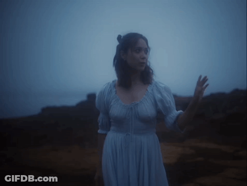 A Woman In A Blue Dress Stands On A Rocky Shore GIF
