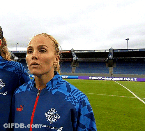 A Woman In A Blue Icelandair Jacket Stands In Front Of A Soccer Field GIF