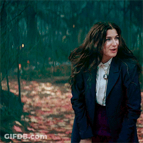A Woman In A Blue Jacket And White Shirt Is Walking Through A Forest GIF
