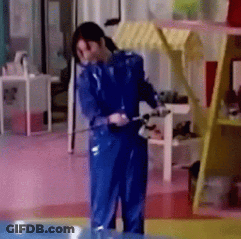 A Woman In A Blue Jumpsuit Is Standing In A Room With A Hose . GIF