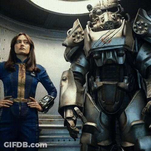 A Woman In A Blue Jumpsuit Stands Next To A Robot With A Red X On Its Chest GIF