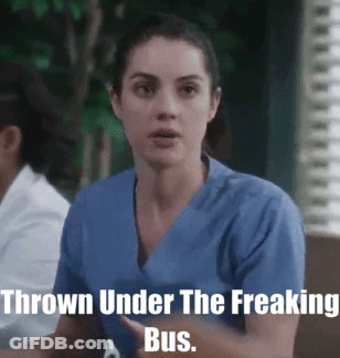 A Woman In A Blue Scrub Is Sitting In Front Of A Sign That Says " Thrown Under The Freaking Bus " GIF