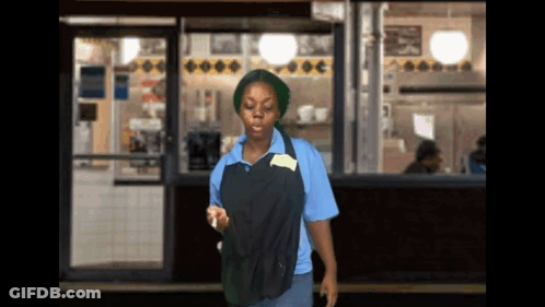 A Woman In A Blue Shirt And Black Apron Is Standing In Front Of A Store GIF