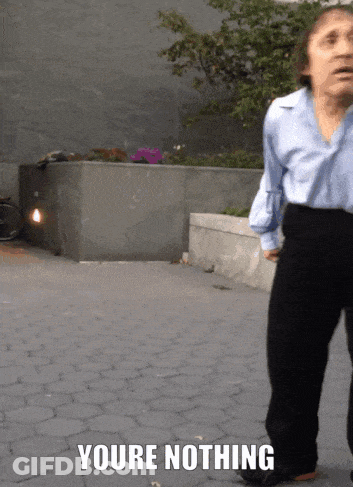 A Woman In A Blue Shirt And Black Pants Is Standing On A Brick Sidewalk With The Words " Youre Nothing " Above Her GIF
