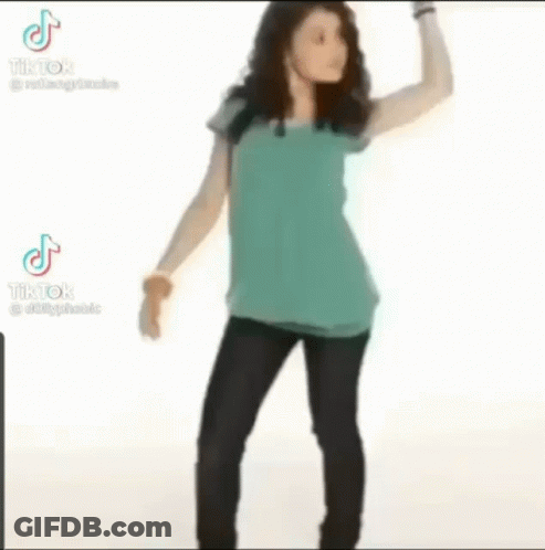 A Woman In A Blue Shirt And Black Pants Is Standing With Her Hands On Her Hips And Looking At The Camera . GIF
