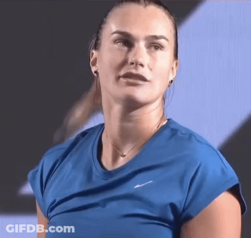 A Woman In A Blue Shirt And Necklace Is Looking At The Camera . GIF