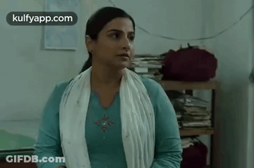 A Woman In A Blue Shirt And White Scarf Is Standing In A Room With A Stack Of Books On A Shelf . GIF