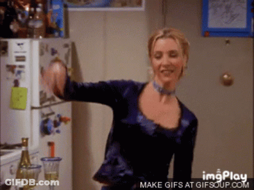 A Woman In A Blue Shirt Is Dancing In Front Of A Refrigerator And A Bottle Of Wine GIF