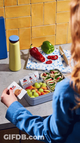 A Woman In A Blue Shirt Is Putting A Sandwich In A Container With Fruit GIF