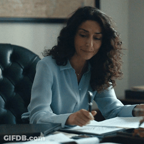 A Woman In A Blue Shirt Is Sitting At A Desk Writing On A Piece Of Paper GIF