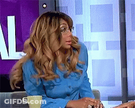 A Woman In A Blue Shirt Is Sitting In Front Of A Purple Wall With The Letter L On It GIF