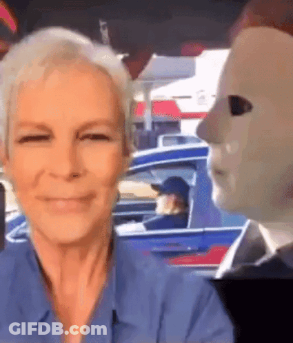 A Woman In A Blue Shirt Is Smiling In Front Of A Man In A Mask . GIF