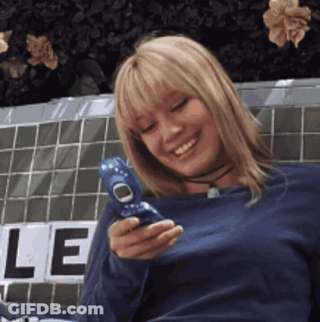 A Woman In A Blue Shirt Is Smiling While Holding A Cell Phone GIF